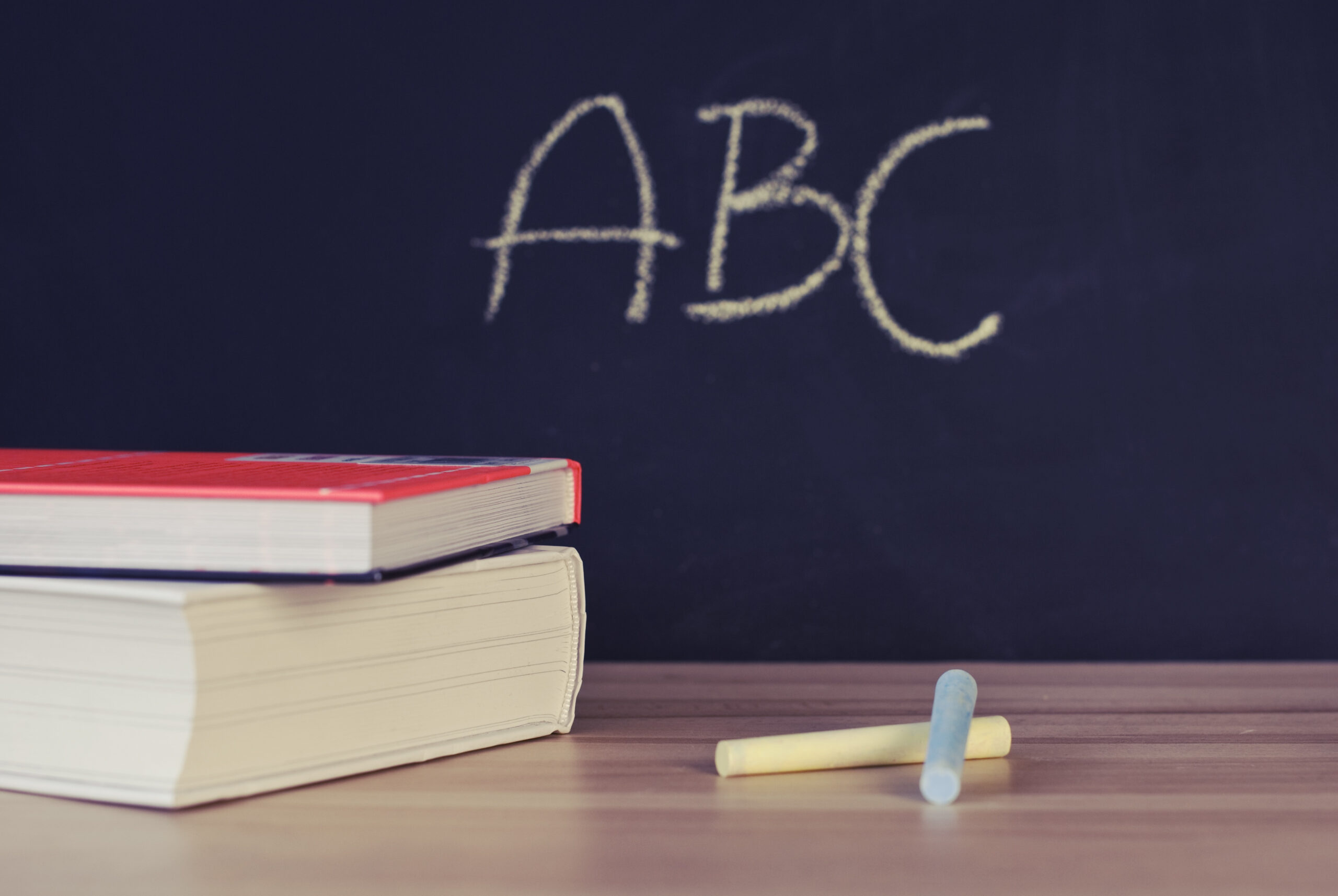 TwoBooks Beside Two Chalks on a desk in front of a blackboard. Om the blackboard are the letters